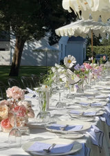 Outdoor wedding reception with tables set for dinner, linen tablecloths and napkins, floral arrangements, and white umbrellas.