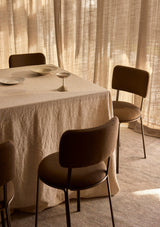 Dining table set with a natural linen tablecloth plates and a candle, surrounded by chairs in a softly lit room.