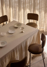 Dining table with natural linen tablecloth and ceramic plates, surrounded by chairs in a softly lit room.